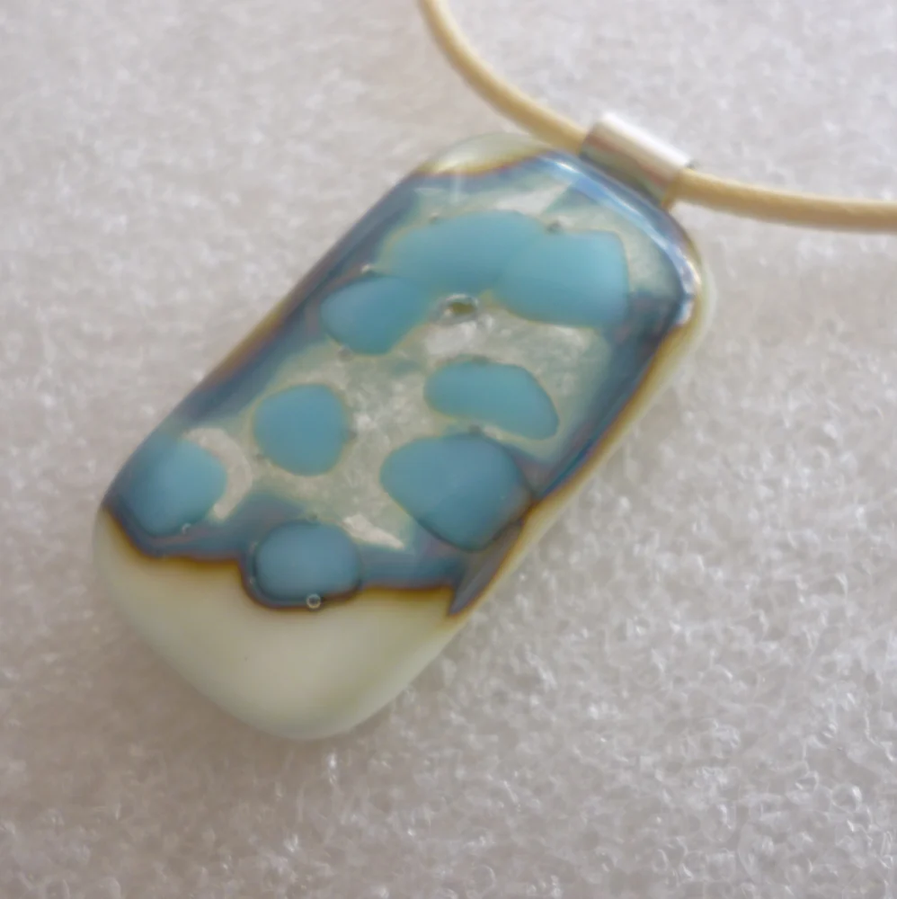 Oblong cream pendant with turquoise, silver and an outline of brown. 925 sterling silver bail attached and hangs from a cream cord necklace.
