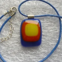 Colourful glass pendant with rounded corners in layers of blue at the bottom, smaller piece of glass in red and smaller piece in yellow. 925 sterling silver bail attached and is hanging from a blue cord necklace with extension chain.