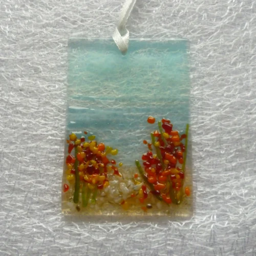 Single seaside suncatcher with a blue background creating the sky and sea, and red and orange flowers with green stems.