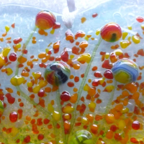 Close up of beads and frit on fused glass heart shaped suncatcher in colours of blue, yellow, green red, orange. This is a very tactile design.