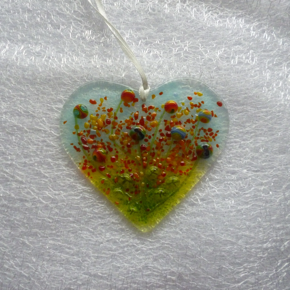 Heart shaped suncatcher with background of green and light blue. Green glass beads for leaves and red, yellow, orange, blue and green colourful glass beads for flowers. White ribbon attached to hang from.