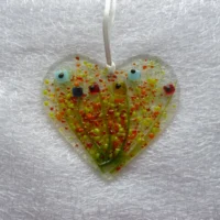 Fused glass heart shaped suncatcher with a floral design. Very colourful with red, blue and yellow quirky square flower heads and long green stems. Green, red and orange speckles of glass as the background and hanging from a white ribbon.