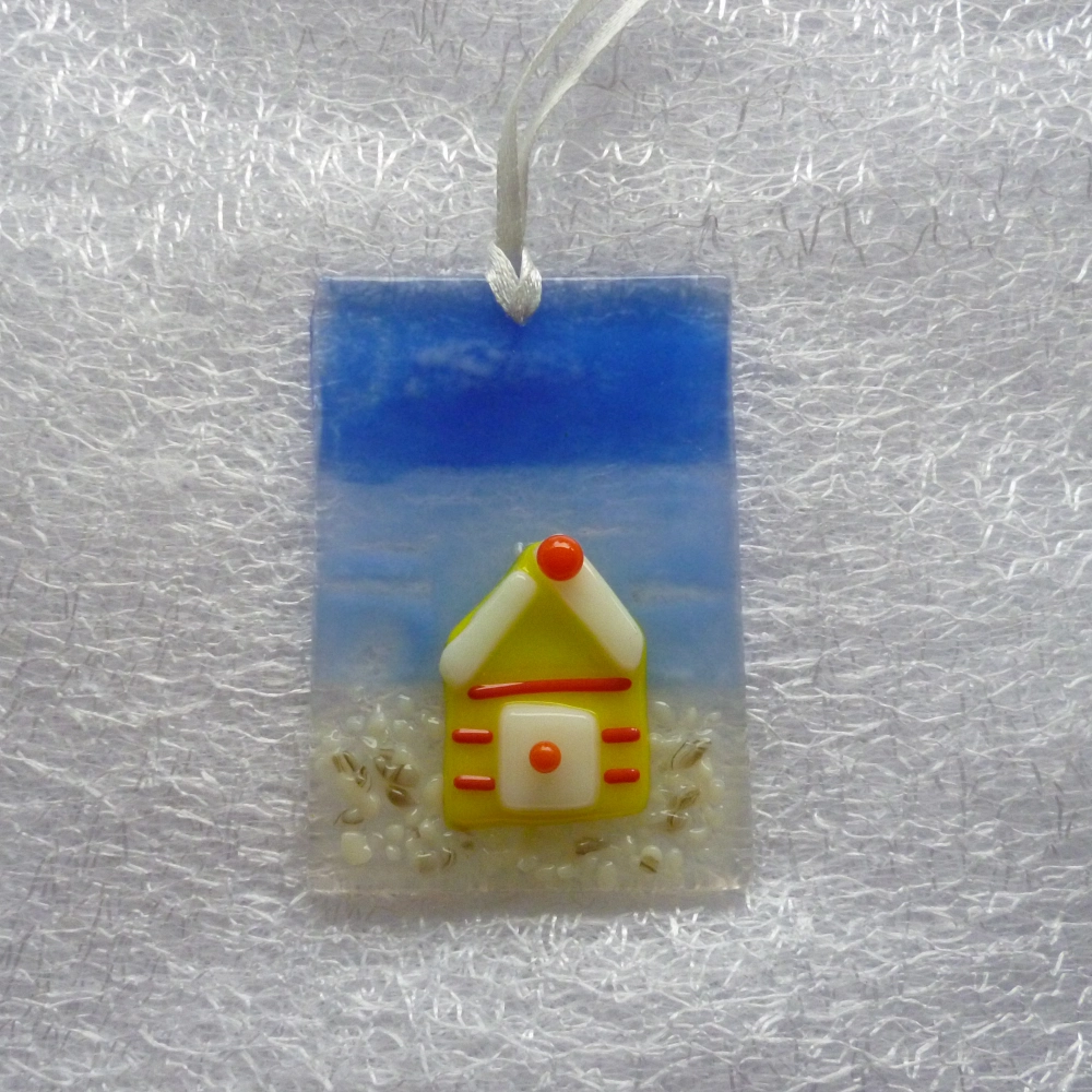 Small suncatcher made with glass, two different shades of blue for the background and a yellow beach hut with red stripes and white door. Hangs from a white ribbon