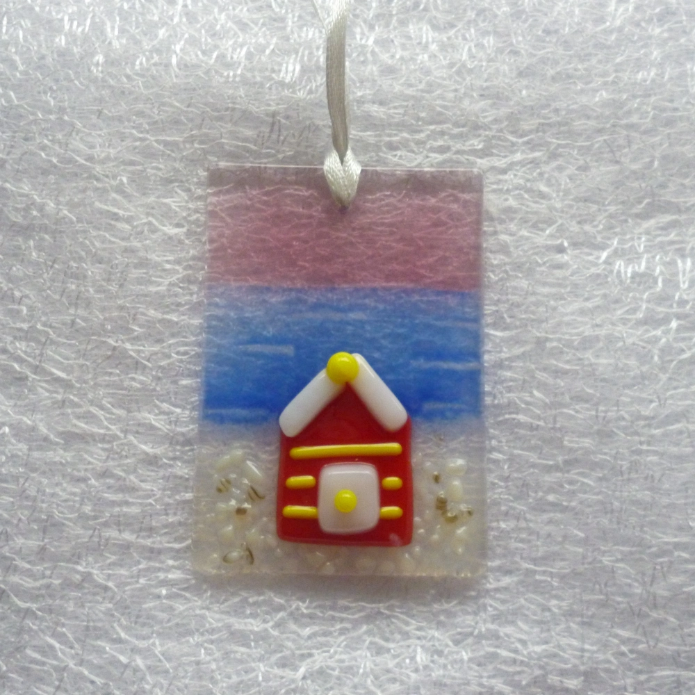 Rectangle shaped fused glass small suncatcher in a coastal design with a background in plum, blue and white. Small glass red beach hut with yellow stripes fused on top. Hangs from a white ribbon.