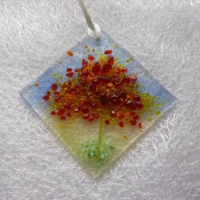 Small suncatcher made with glass. A diamond shaped fused glass suncatcher with blue and green background. The tree is very tactile in colours of red, orange, yellow and green in the design of a tree.