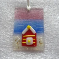 Small suncatcher hanging from a white ribbon. Rectangle in shape with a background of plum blue and white. Beach hut in red glass with yellow stripes.