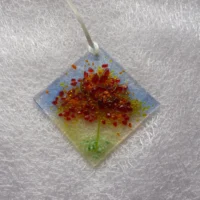 Small suncatcher made of fused glass with the design of an autumn tree, and colours of red, orange and yellow for the leaves of the tree and a green stringers for the trunk. The suncatcher hangs from a white ribbon.