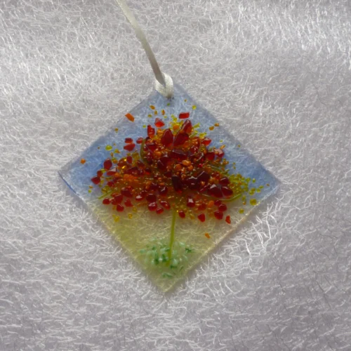 Small suncatcher made of fused glass with the design of an autumn tree, and colours of red, orange and yellow for the leaves of the tree and a green stringers for the trunk. The suncatcher hangs from a white ribbon.