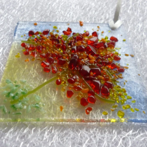 Colours of red, yellow, orange and green frit fused onto a triangular piece of glass in a design of an autumn tree. White ribbon at the top to hang from. A very tactile design.