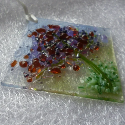 Small suncatcher made of glass with a light blue and green background. Stringer to represent the trunk of a tree with blossoms in plum, lilac and purple.