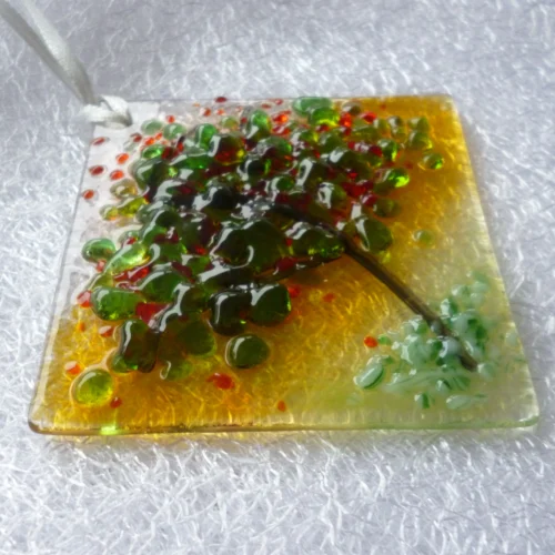Side view of small suncatcher in a tree design, with a background of orange and green. Stringer has been used as the trunk of the tree with green and red frit for the leaves and blossom.