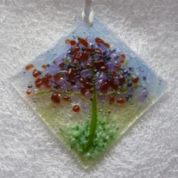 Small suncatcher made with glass. A diamond shaped fused glass suncatcher with pale blue and green background. The tree is very tactile in colours of plum, purple, green and lilac.