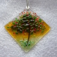 Small suncatcher made of fused glass with the design of a summer tree, and colours of red and green frit fused on top. This suncatcher hangs from a white ribbon.