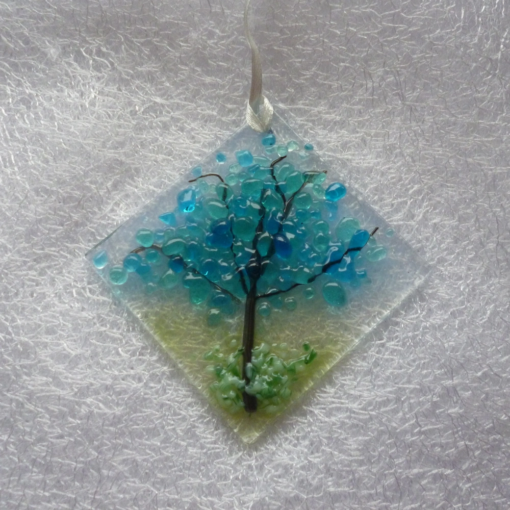 diamond shaped fused glass winter tree small suncatcher. Blue and turquoise frit creates the blossoms with black trunk and branches with green and white at the bottom for the grass. Hole at the top with a white ribbon threaded through to hang from.