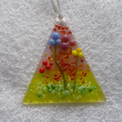 triangular fused glass suncatcher with flowers made of glass beads in blue, plum, red and yellow with green stems. Background of green and pink.