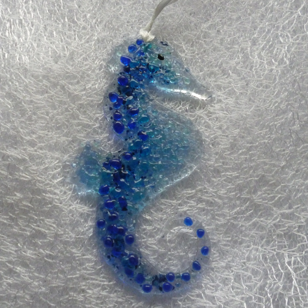 A very tactile fused glass seahorse using blue and turquoise frit. Hanging from white ribbon.