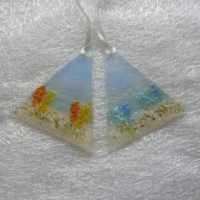 Two small triangular coastal suncatchers, with a background in transparent blue and cream. One suncatcher has blue flowers and the other with orange flowers. Hangs from a white ribbon.