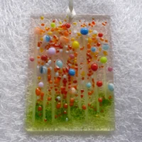 Fused glass suncatcher with colourful beads to create flowers in blue, orange, yellow, green, red and pink. Red and orange frit used for decoration and hangs from a white ribbon.
