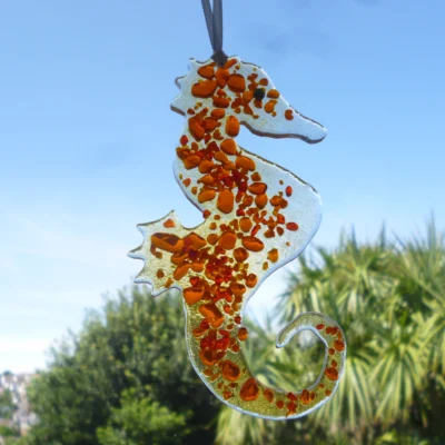 Orange and clear glass seahorse hanging outside with trees in the background.