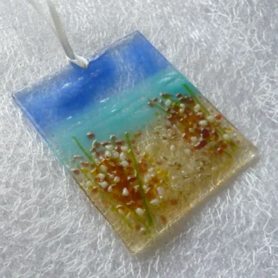 Side view of coastal suncatcher with plum and white flowers with green stems. Background in blue turquoise and tan, with a ribbon threaded through the top.