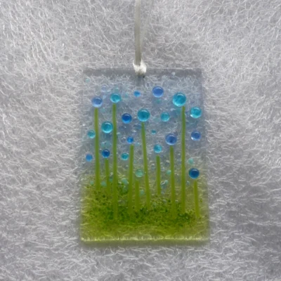 Glass suncatcher with blue and turquoise beads as flowers and green stringers for stems. Hole at the top with a white ribbon threaded through.