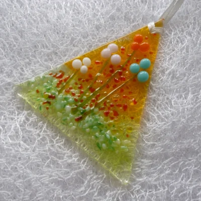 triangular glass suncatcher with flowers in orange, pink, white and blue on a background of green and orange. Hanging from a white ribbon.