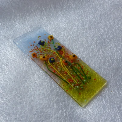 Side view of tactile blue and orange flower suncatcher. Background colours of blue, pink and green with quirky square glass flower and long stems. Hanging from a clear cord.