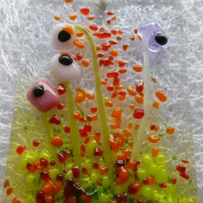 Close up of pink and lilac flower suncatcher, with each flower having a black centre with green stems and red, orange and green frit detailing.