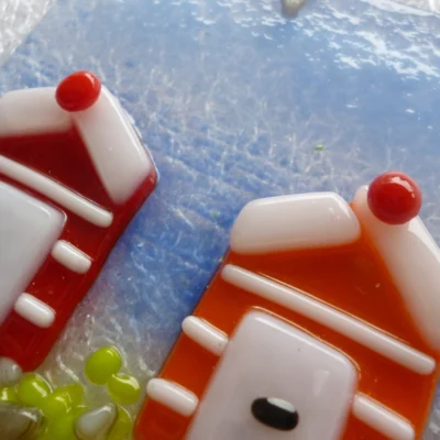 Close up of red and orange beach huts on coastal suncatcher. Background of suncatcher is blue, and the huts have white stripes.