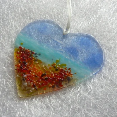 Side view of heart shaped glass suncatcher, in colours of blue, turquoise, tank, red, orange and yellow. Hanging from a white ribbon.