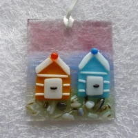 Rectangle shaped coastal suncatcher with tactile beach huts in orange and blue. Background of suncatcher is pink and blue and hangs from a white ribbon.