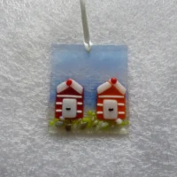 Rectangle shaped coastal suncatcher with red and orange beach huts with white stripes. Large frit at the bottom of the suncatcher to create stones. Hanging from a white ribbon.