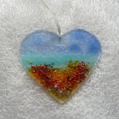 Fused glass heart suncatcher. Background in blue, turquoise and tan, with frit in colours of red, orange and yellow. Hanging from a white ribbon.