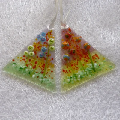 Two triangle shaped fused glass suncatchers hanging from white ribbon. One suncatcher has multicoloured beads to create flowers with stems and the other has green heart and blue hearts. Red and green frit for decoration.
