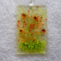 Brightly coloured orange flower suncatcher, with a background in a very pale orange. Tactile glass flowers in a bright orange with green stems and green frit at the bottom to represent grass. Red and orange frit used for added detail. Hangs from a white ribbon.