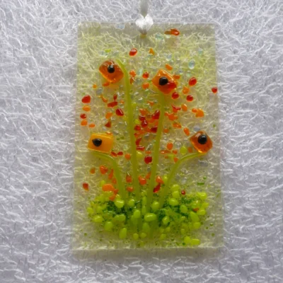 Brightly coloured orange flower suncatcher, with a background in a very pale orange. Tactile glass flowers in a bright orange with green stems and green frit at the bottom to represent grass. Red and orange frit used for added detail. Hangs from a white ribbon.