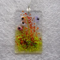 Rectangle fused glass flower suncatcher. Quirky square shaped pink and lilac flowers with black centres, red, orange and green frit for decoration. Hangs from a white ribbon.