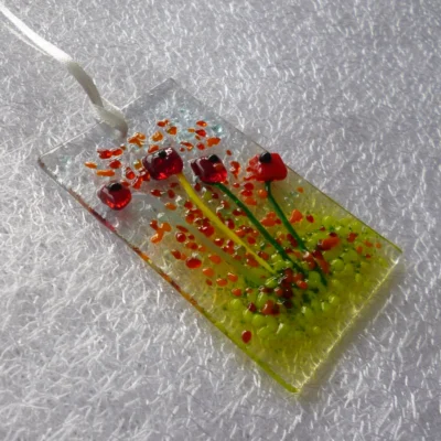 Side view of tactile red flower suncatcher with red, green and orange frit for decoration. Hole at the top with a white ribbon threaded through for display.