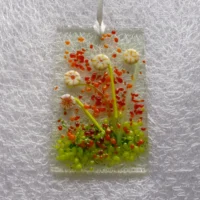 Small rectangle fused glass cream flower suncatcher with red, orange and green frit for decoration. Hangs from a white ribbon.