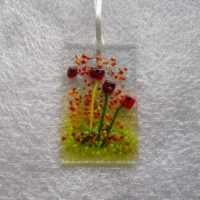Fused glass red flower suncatcher hanging from a white ribbon. Red, orange and green frit for decoration.