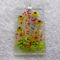 Rectangle fused glass yellow flower suncatcher. Suncatcher has four quirky flowers with black centres, and frit decoration in vibrant red, orange and green. Hangs from a white ribbon.