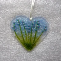 Fused glass heart suncatcher with blue flowers and green stems, blue frit (crushed glass) for decoration and hangs from a white ribbon.
