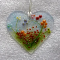 Pretty glass heart suncatcher with vibrant floral design. Background of blue and green with red frit as decoration. Hanging from a ribbon.