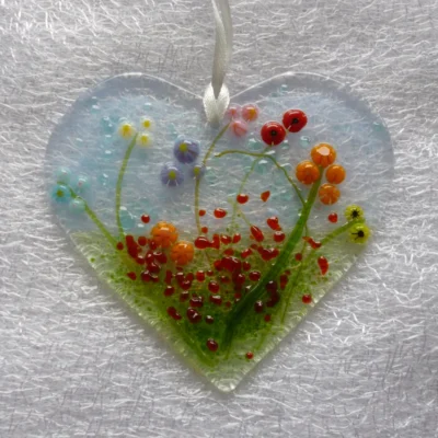 Pretty glass heart suncatcher with vibrant floral design. Background of blue and green with red frit as decoration. Hanging from a ribbon.