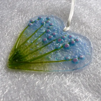 side view of fused glass heart suncatcher with blue and pink flowers with long green stems. Hangs from a white ribbon.
