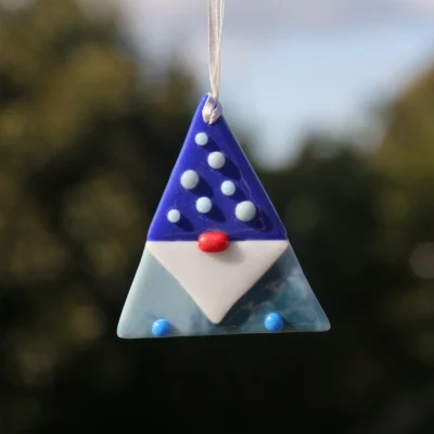 Fused glass triangle shaped gonk with light blue body, dark blue hat with small beads on top, white beard, red nose, and hanging from a white ribbon.
