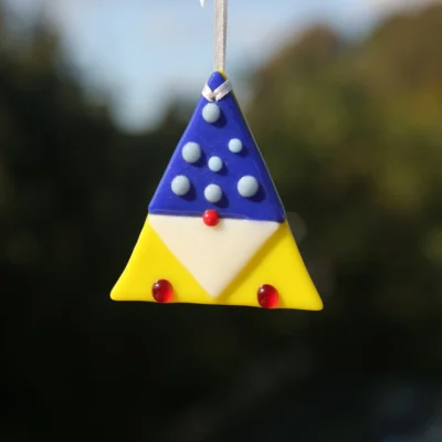 Vibrant fused glass triangle shaped gonk with a yellow body, dark blue hat with small beads on top, white beard, red nose, and hanging from a white ribbon.