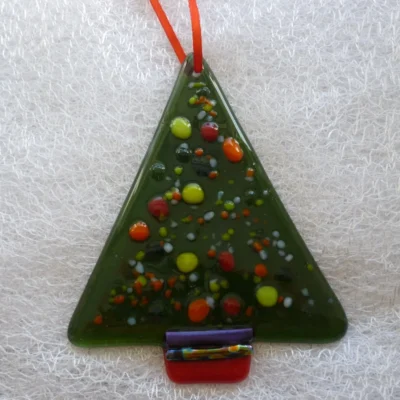 Fused glass triangle shaped Christmas tree with frit for decoration laying on a white cloth, hanging from a red ribbon.