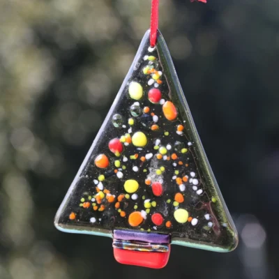 Triangle shaped green transparent fused glass Christmas tree, with colourful frit in yellow, orange, red and white. Dichroic glass at the bottom of the tree along with red glass to represent the trunk.