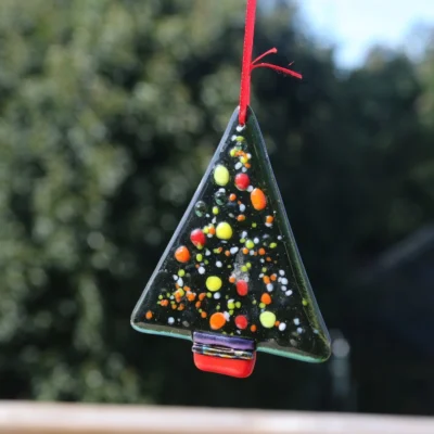 Green fused glass Christmas tree with colourful frit in red, yellow, orange and white, hang in a window from a red ribbon.